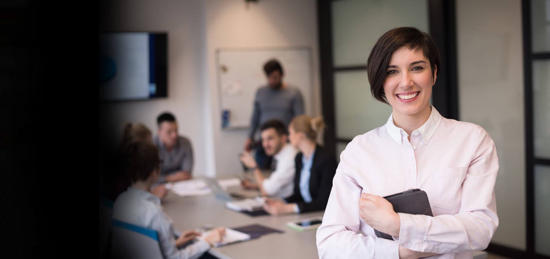 Confident woman in a white shirt stands in a meeting room, holding a tablet
