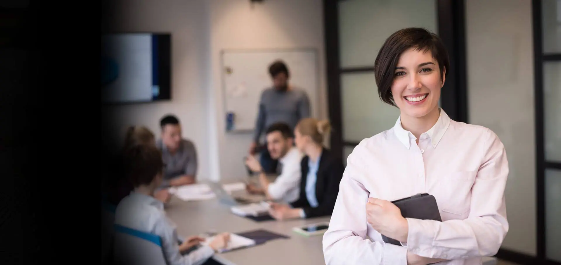 Confident woman in a white shirt stands in a meeting room, holding a tablet