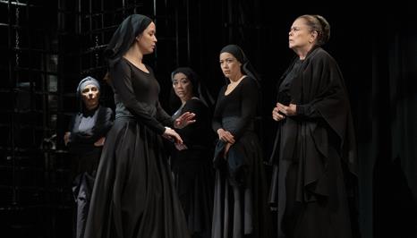 A group of five women in black costumes stands in a theatrical setting, engaged in a tense conversation, conveying themes of drama and conflict.